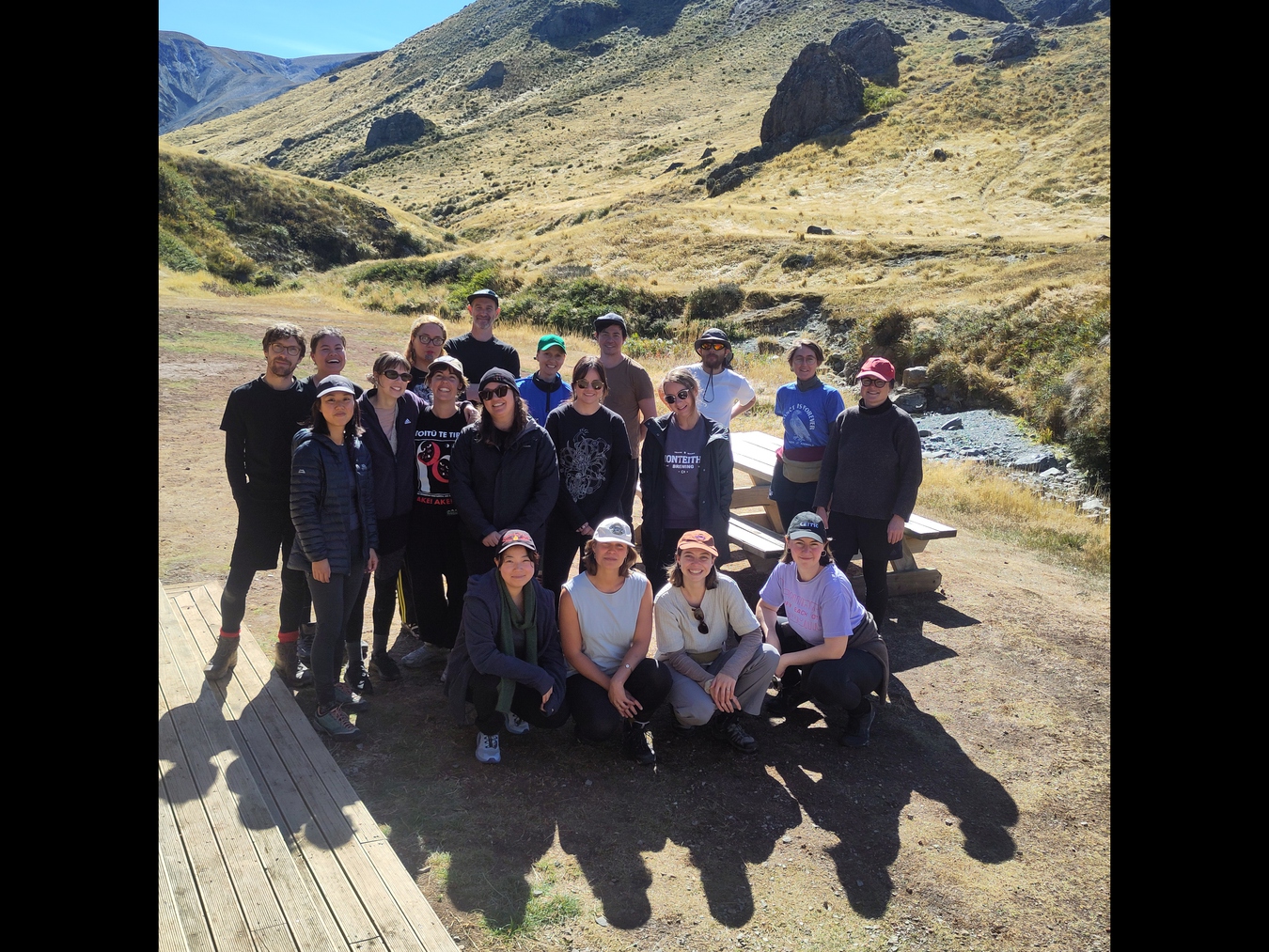 A group photo is taken outside the hut before we leave on the second day. The sky is blue and a rocky peak can be seen high behind us